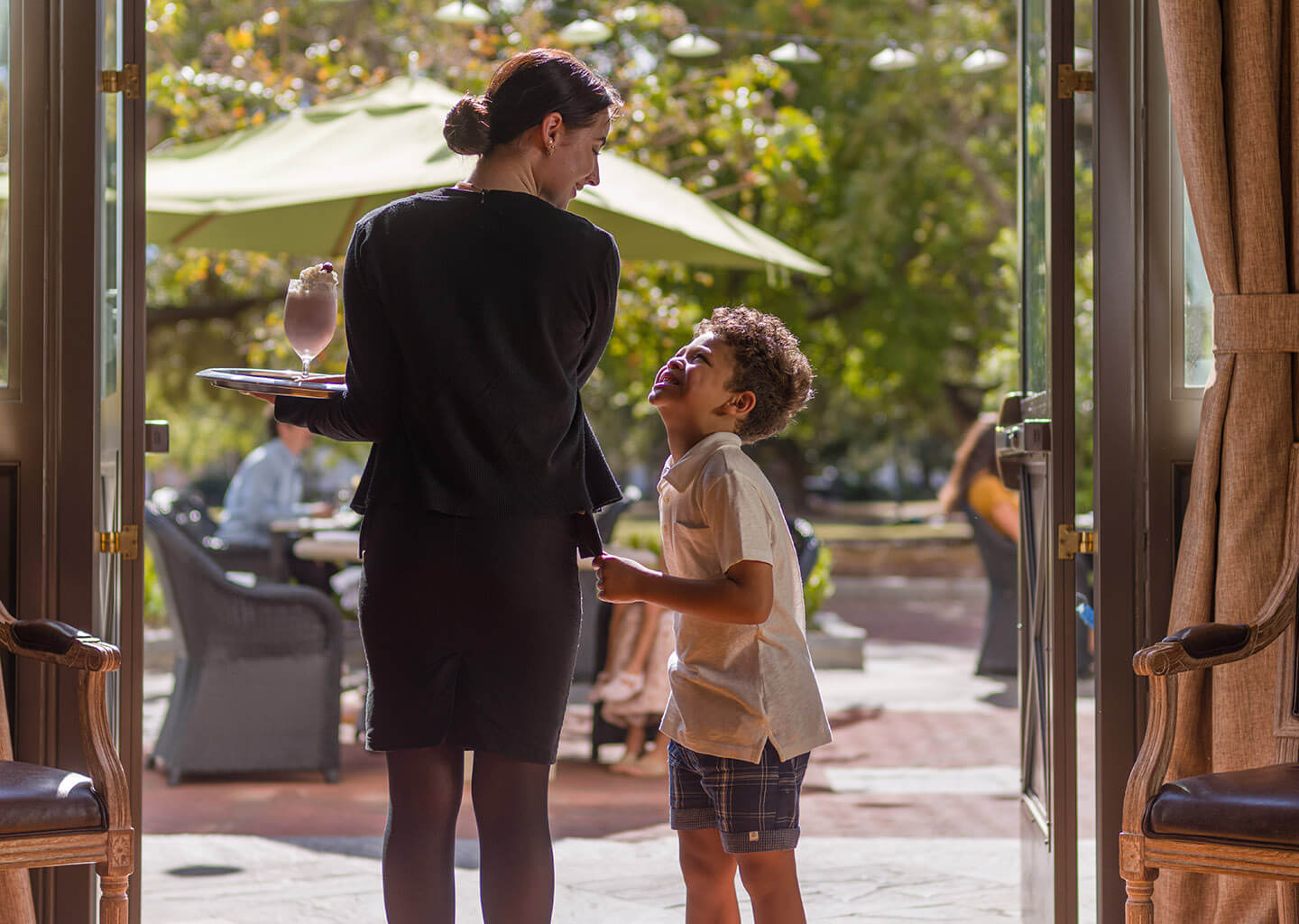 Lady holding a strawberry milkshake on a tray smiling at a child on Gabrielle's terrace